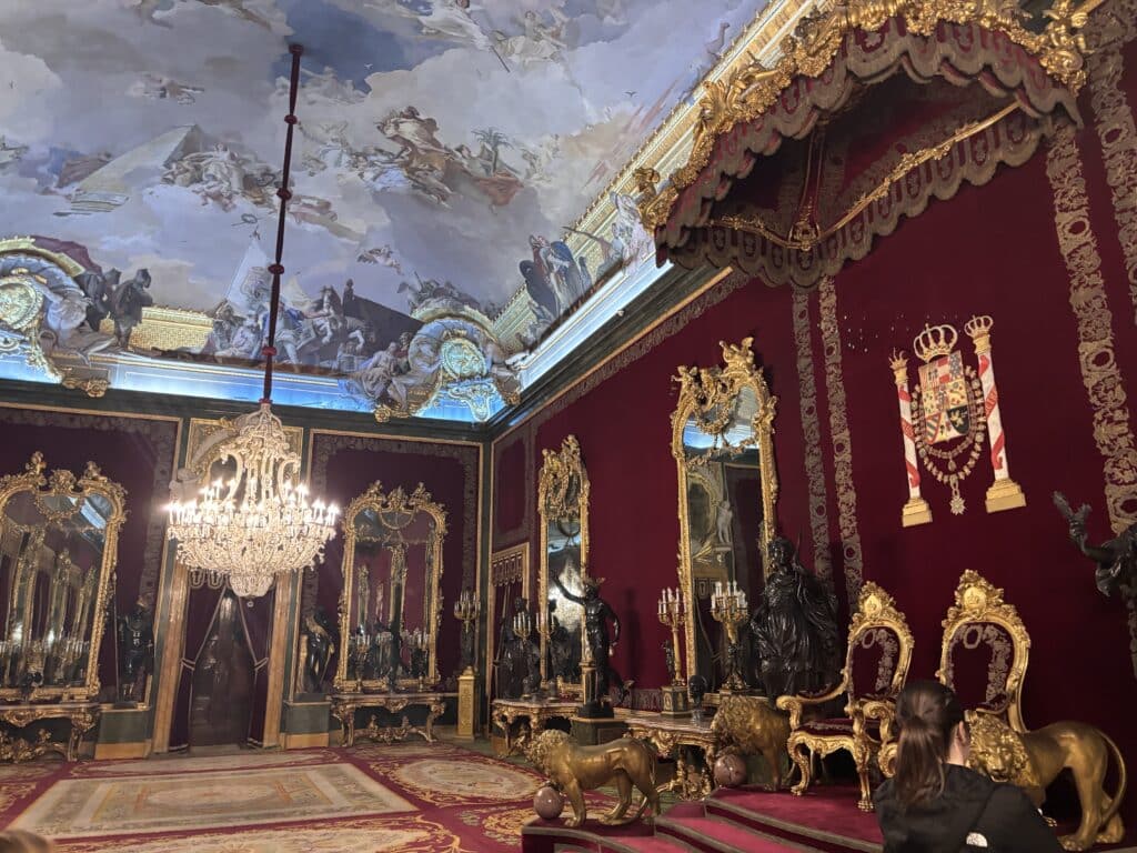 Ornate throne room with red velvet walls, gold carvings, and a large gilded throne; regal decor and chandeliers visible.