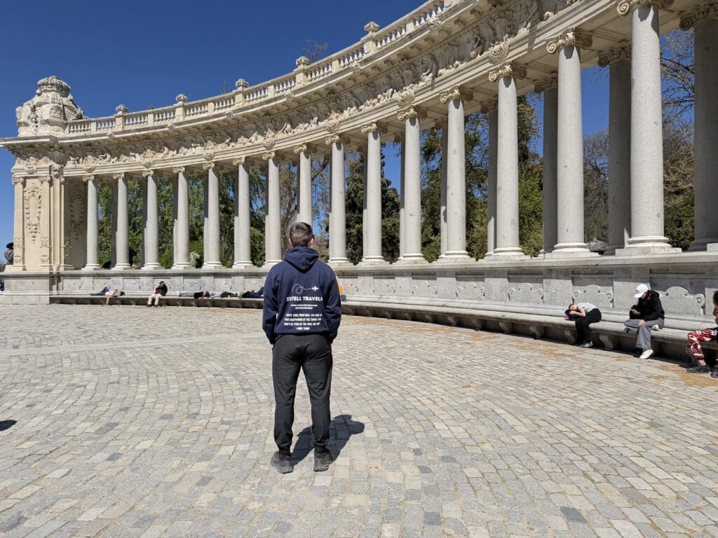 Man in a navy hoodie stands with his back to the camera in front of a curved, ornate stone colonnade under a clear blue sky.