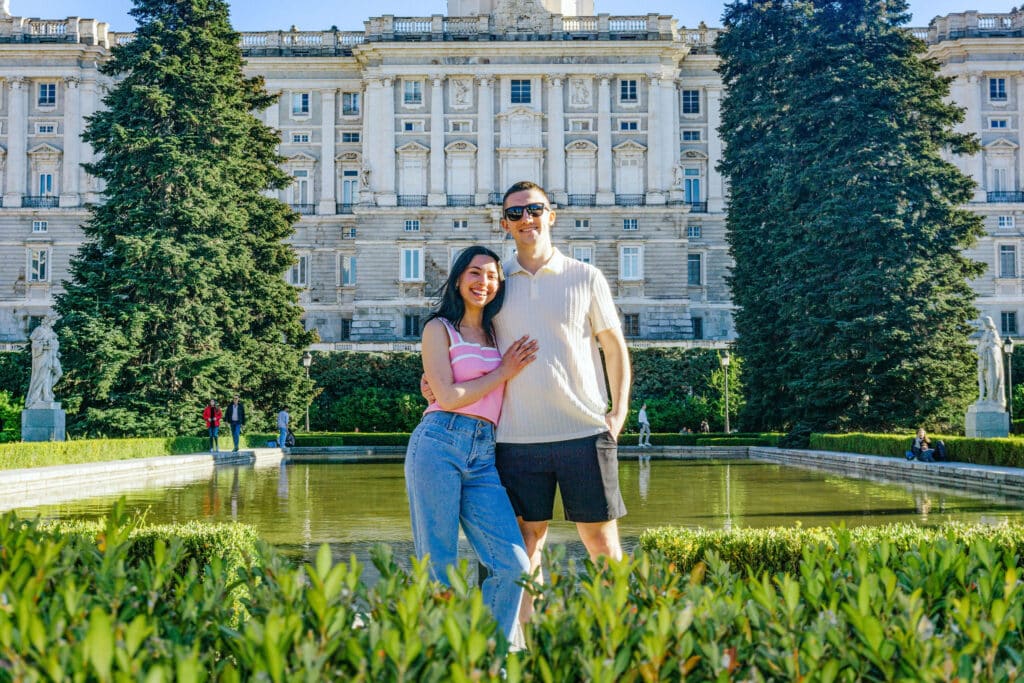 Smiling couple posing in front of a grand white neoclassical building with statues and a reflecting pool in the foreground.