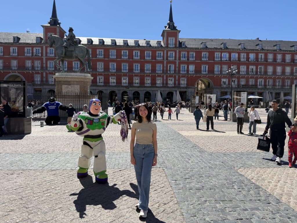 A young woman smiles at the camera in a sunny cobblestone plaza, with a large equestrian statue and a red historic building façade in the background.