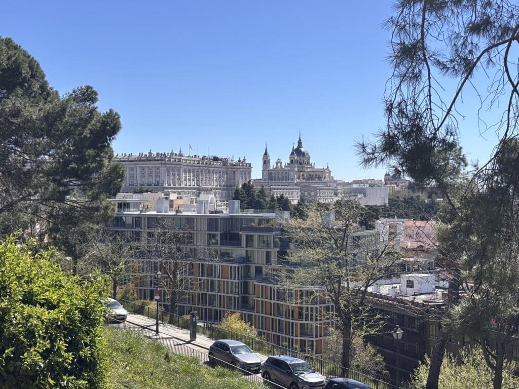 Cityscape with modern glass apartment block in the foreground and a historic domed church in the distance, framed by green trees on a sunny day.