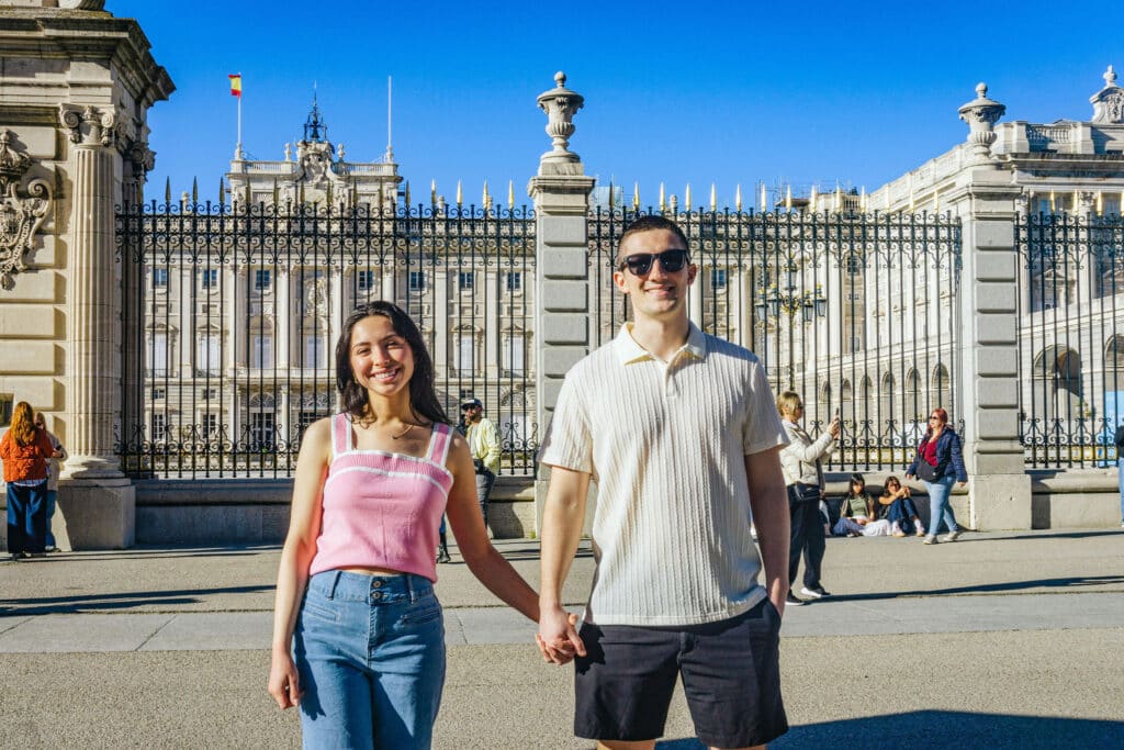 Two young adults holding hands and smiling in front of an ornate iron fence at a grand historic building on a sunny day.