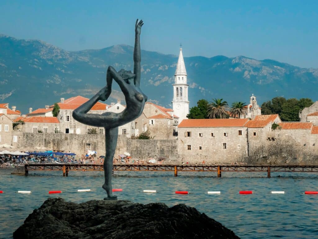 Bronze statue of a dancer balancing on one leg with arms raised, set in a harbor with rocky foreground and red-roofed stone buildings and a church tower in the background against mountains and blue sky.