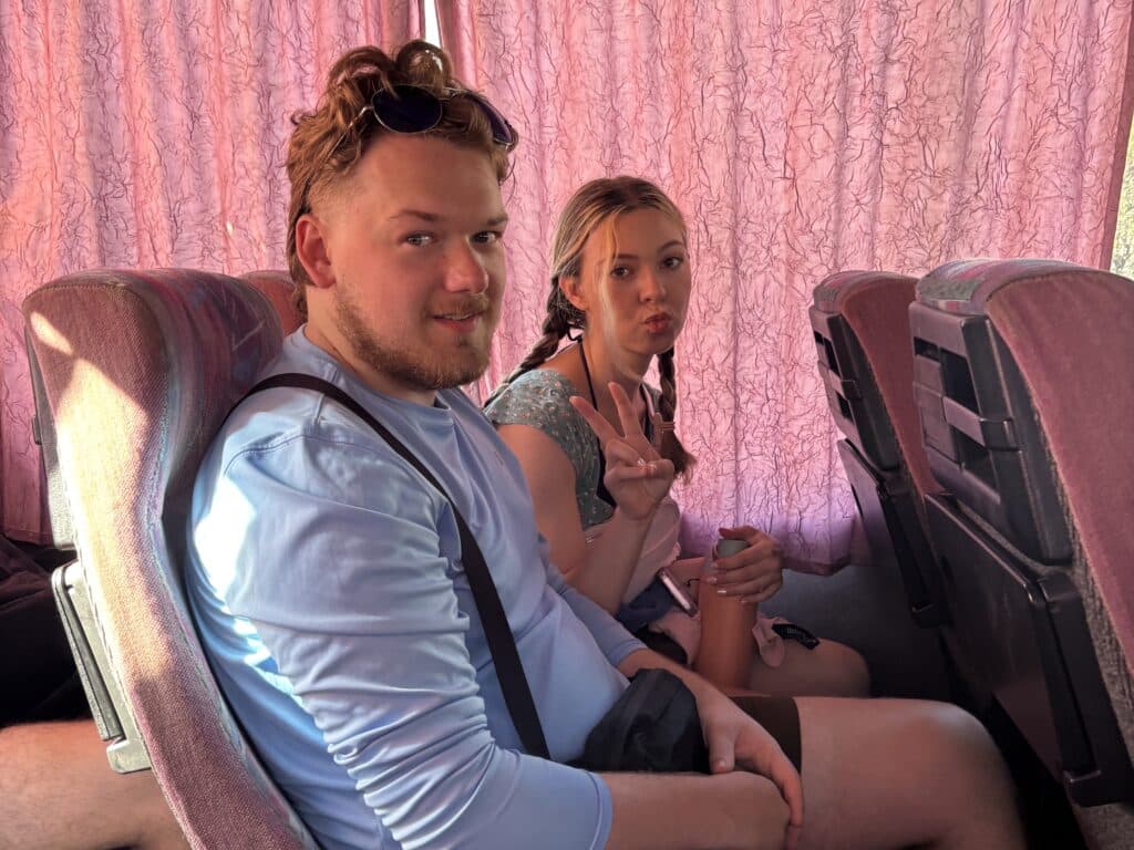 Two people sit on a bus with pink upholstery, posing for a photo; man in a blue shirt and sunglasses on his head, woman with braids making a peace sign and puckering her lips.