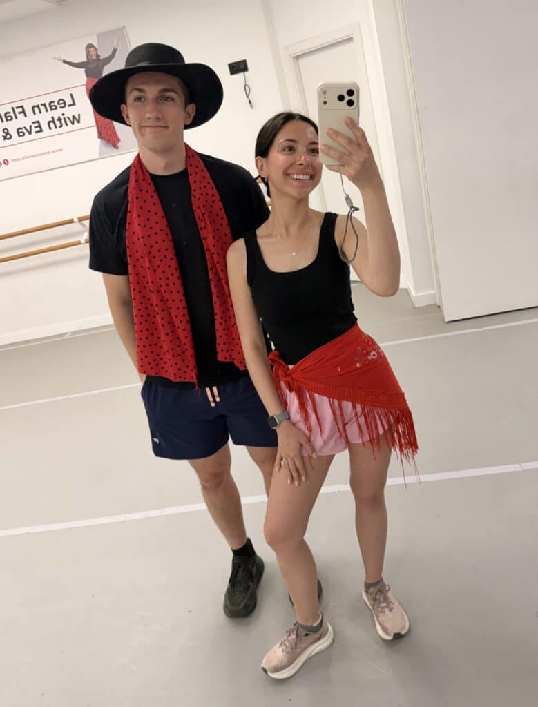 Two dancers take a mirror selfie in a studio; she wears a black tank top and pink shorts with a red fringed wrap, he wears a black shirt, black hat, and red scarf with dots.