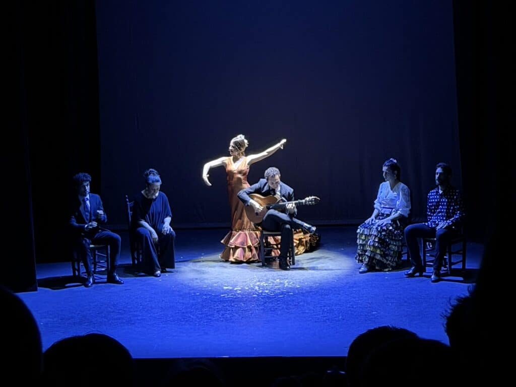 Stage performance: a dancer in an orange dress raises one arm beside a guitarist; seated musicians flank them under blue lighting.