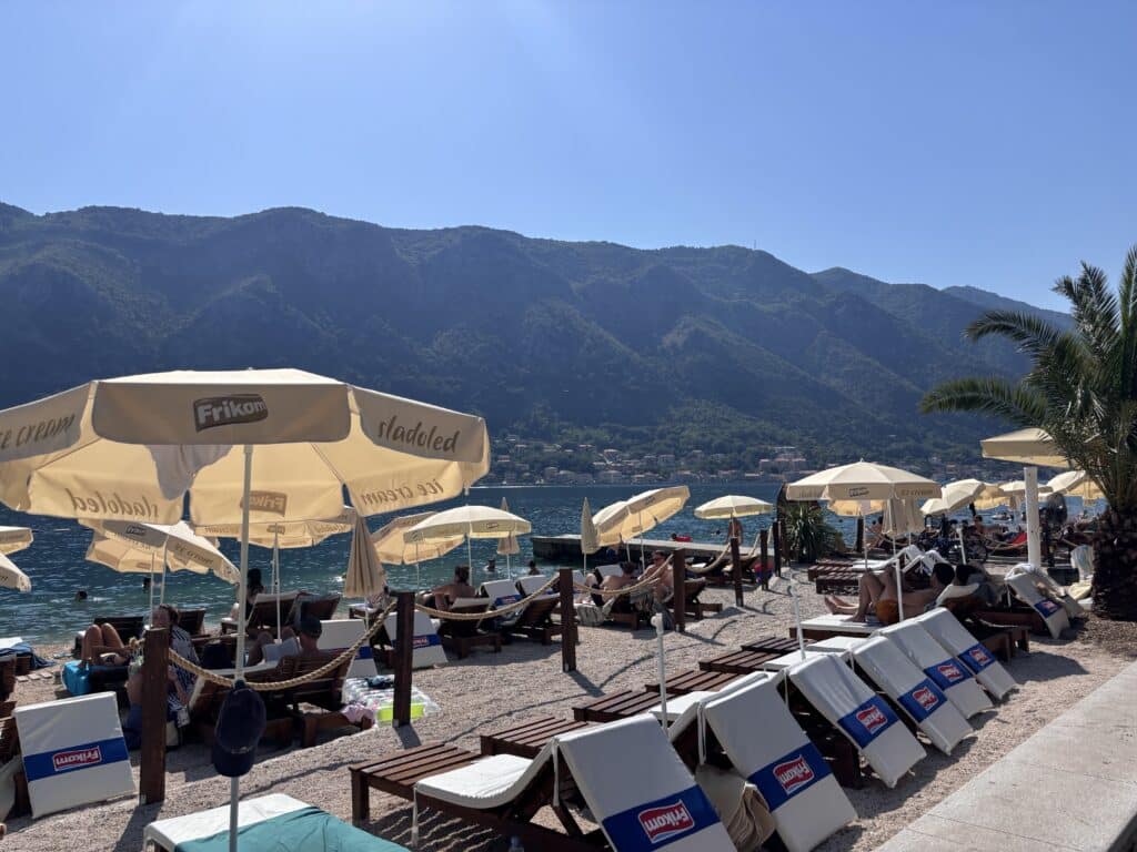 Busy beach with beige umbrellas and sun loungers by the blue water; green mountains in the distance; clear sky above.