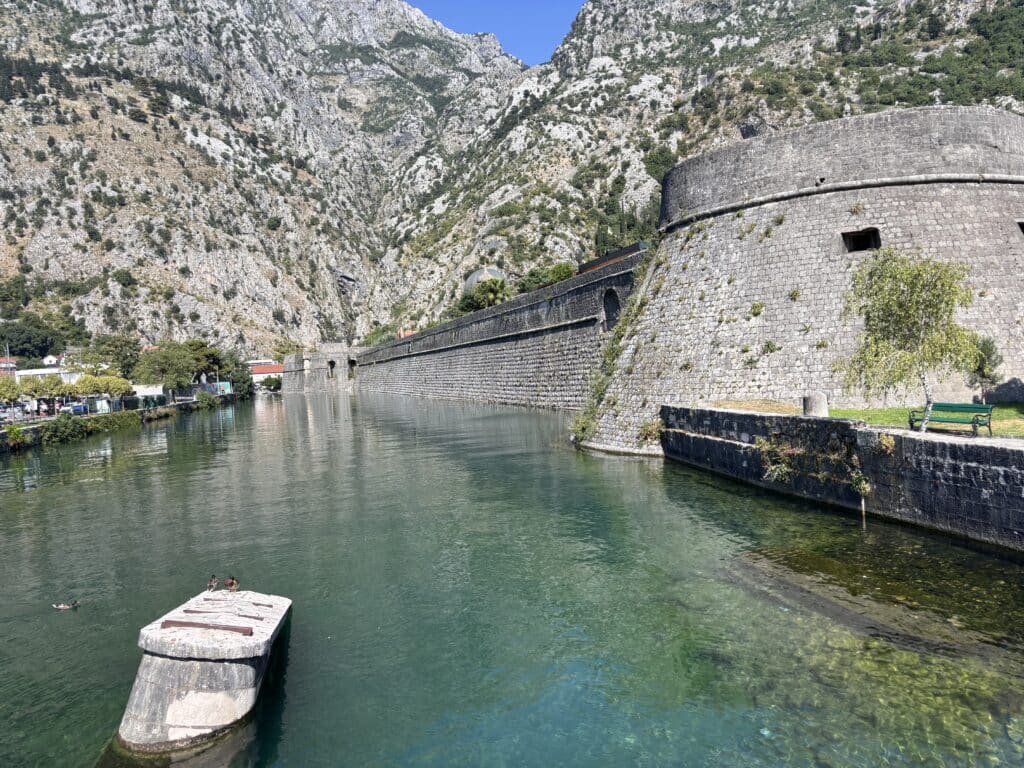 Stone fortress wall along a calm river with steep rocky mountains in the background.