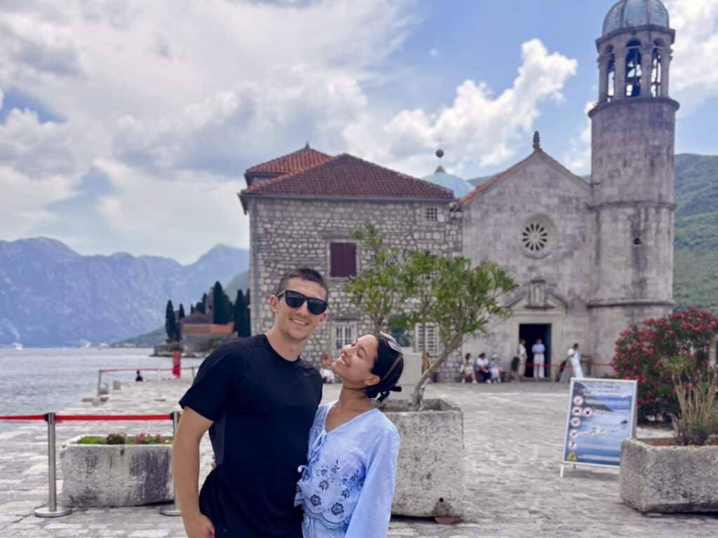 Couple posing and smiling in front of a stone church by the sea, with mountains in the background.