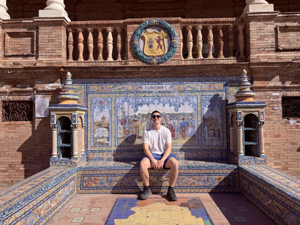 Man wearing a gray shirt and shorts sits on a colorful tiled bench in front of a brick courtyard with ceramic mosaics and crest.
