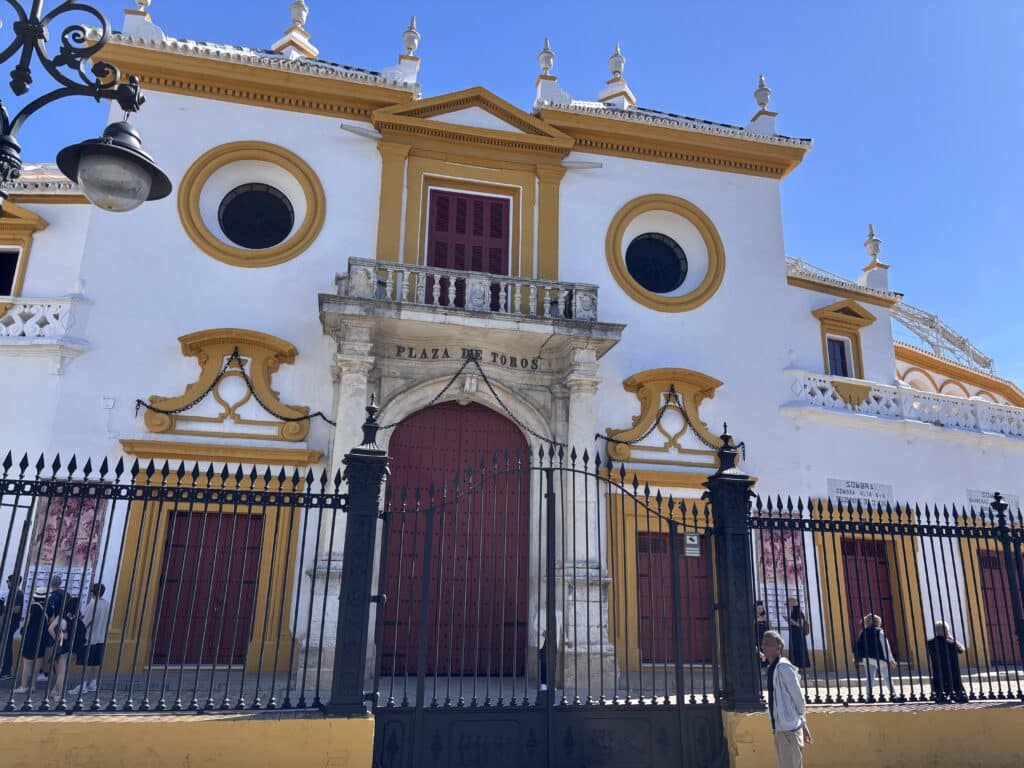 Facade of a bullring with white walls and yellow trim, circular windows, a central balcony, and a red door labeled Plaza de Toros, behind a black iron fence.
