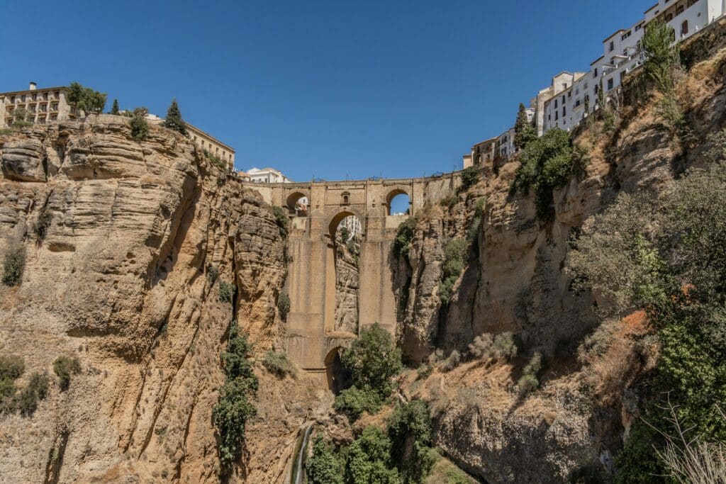 Historic stone bridge with arched spans across a deep rocky gorge, white buildings atop the cliffs and clear blue sky above.