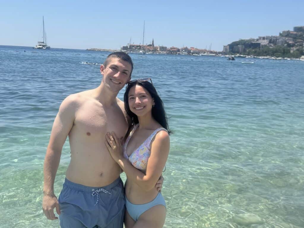 Smiling couple stands in shallow turquoise water at the beach, with a coastal town and boats in the background.